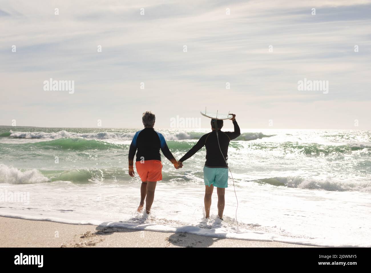 Rear view of biracial senior man carrying surfboard on head holding hand of woman walking at beach Stock Photo