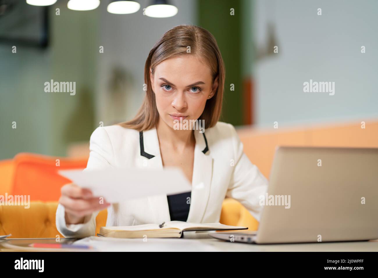 Business Woman in a White Elegant Jacket Sits at her Desk with a Laptop, Gazes to the Camera and Gives Business Papers. Blonde M Stock Photo