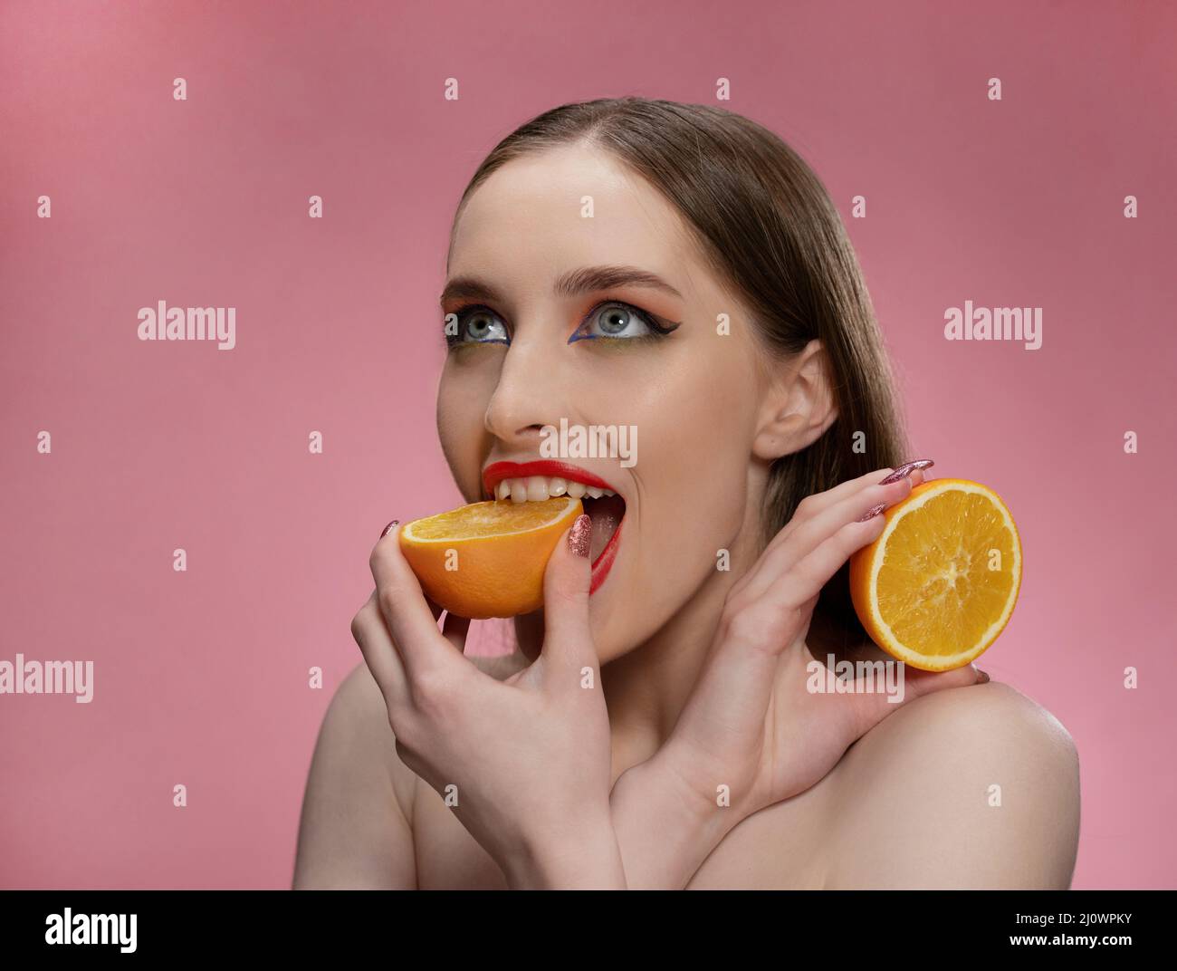 Beautiful fashion girl holding orange sliced in half in her hand biting it looking at camera. Charming joyful funny lady with re Stock Photo