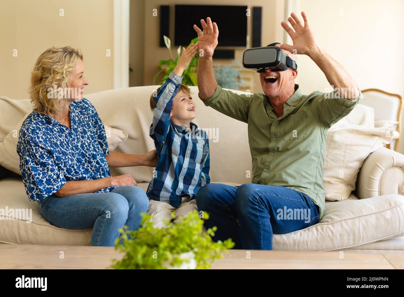 Happy caucasian grandmother and grandson watching grandfather wearing vr headset on couch at home Stock Photo