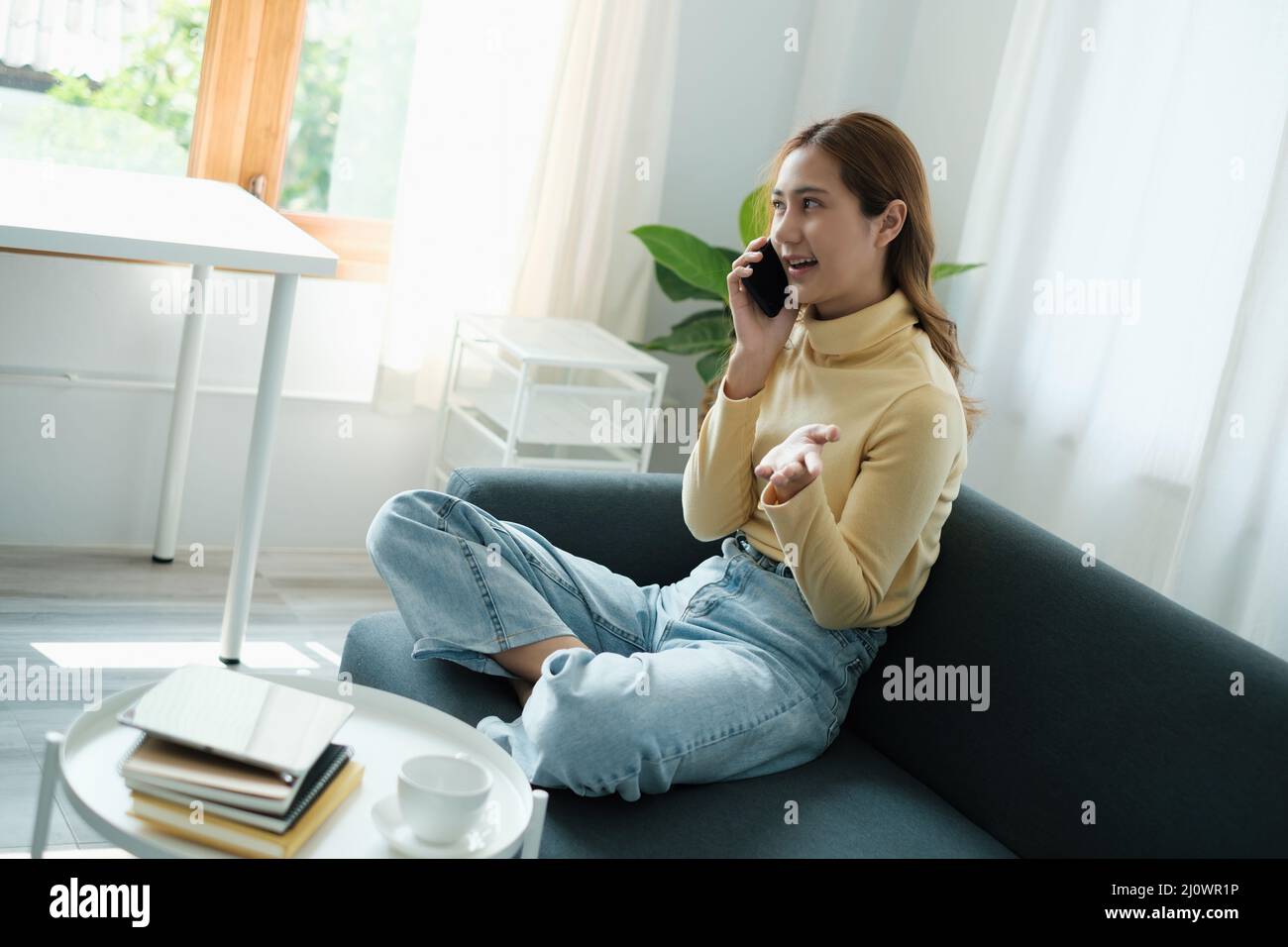 Young Asian woman talking with friend by cell phone while sitting on sofa at home. Stock Photo