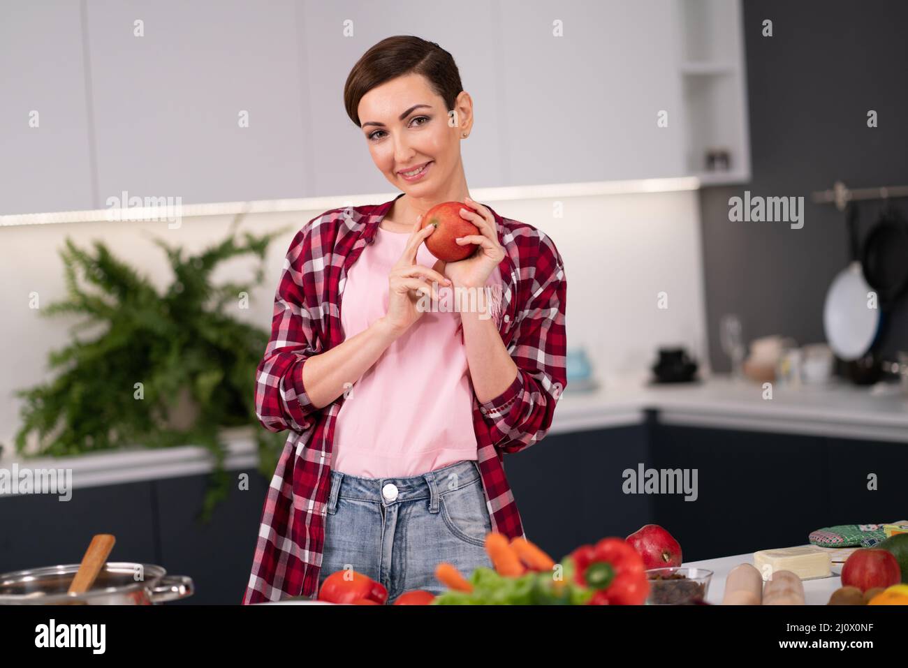 Happy young woman with apple in hand standing in the kitchen. Short hair woman in red plaid shirt in the modern kitchen. Nutriti Stock Photo