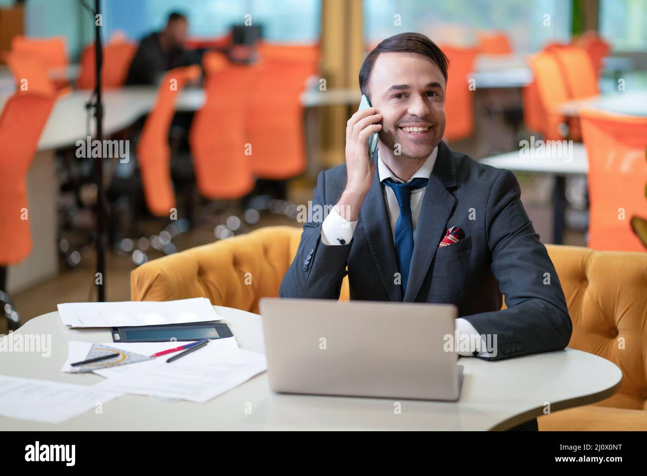 Smiling young businessman or manager talking on the phone while working on laptop sitting at the modern office with toxic orange Stock Photo