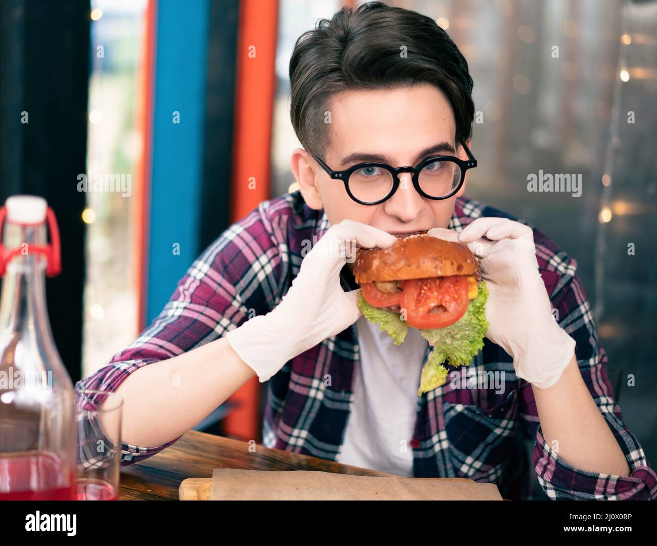 Teenager boy eats falling apart burger sitting at restaurant terrace. Handsome young man in glasses wearing rubber glows holding Stock Photo