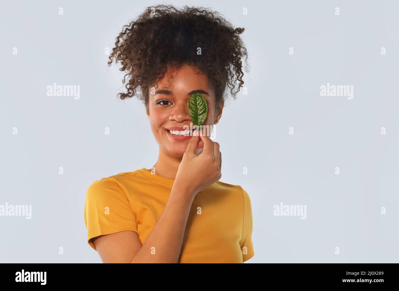 Happy Afro american girl in yellow tshirt with curly hair covering one eye with green leaf Stock Photo