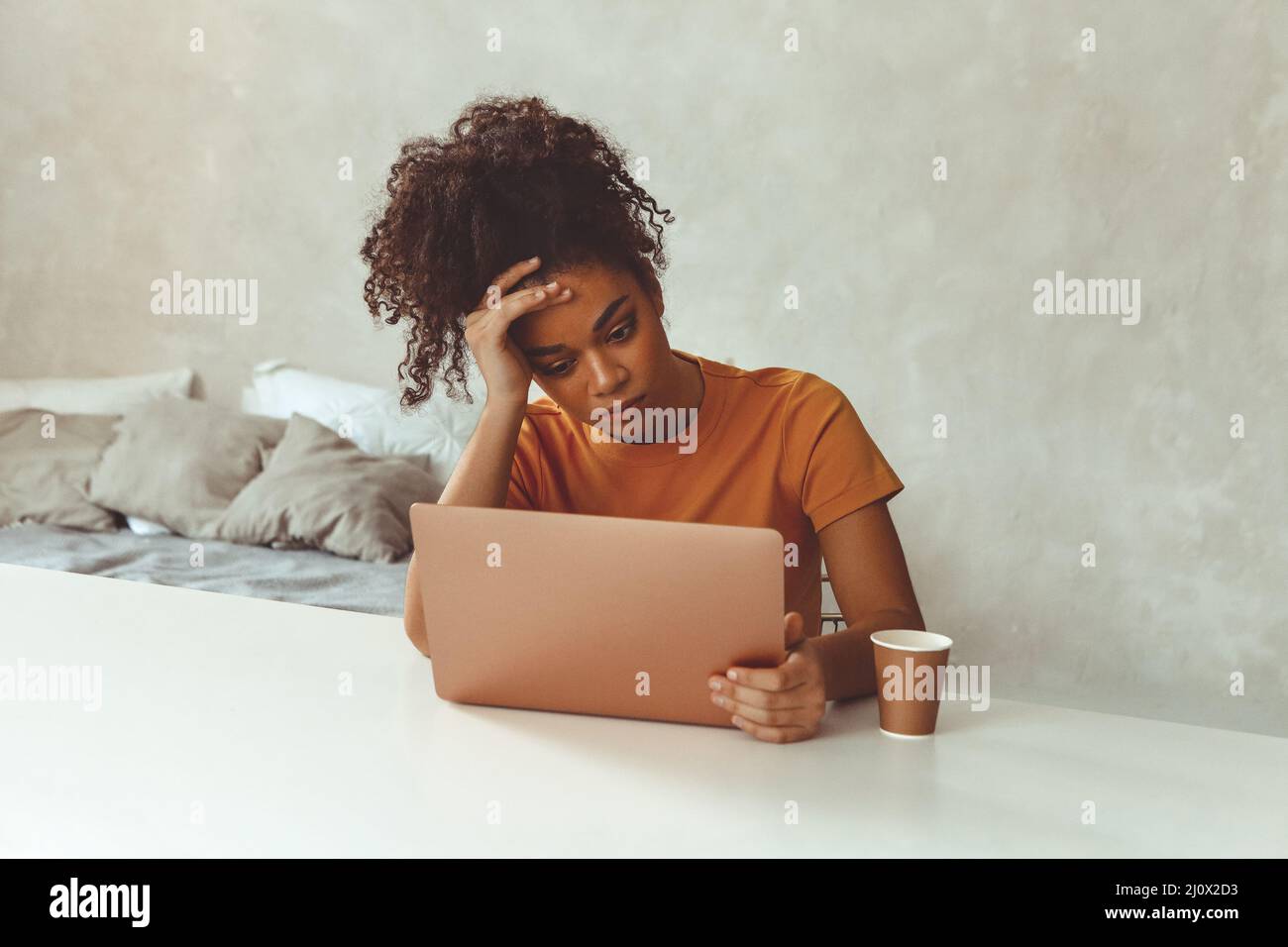 Tired flustrated African descent young girl sitting at desk in front of laptop while irritably looking at computer screen with h Stock Photo