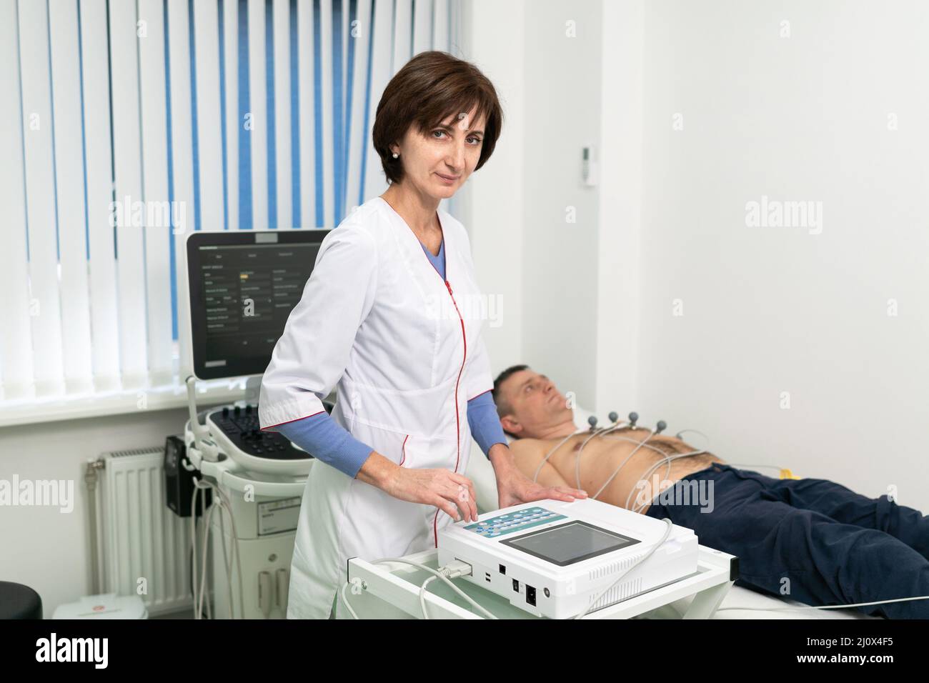 Cardiologist doctor performs electrocardiography procedure on man lying on couch in hospital. ECG test of male at modern clinic. Stock Photo