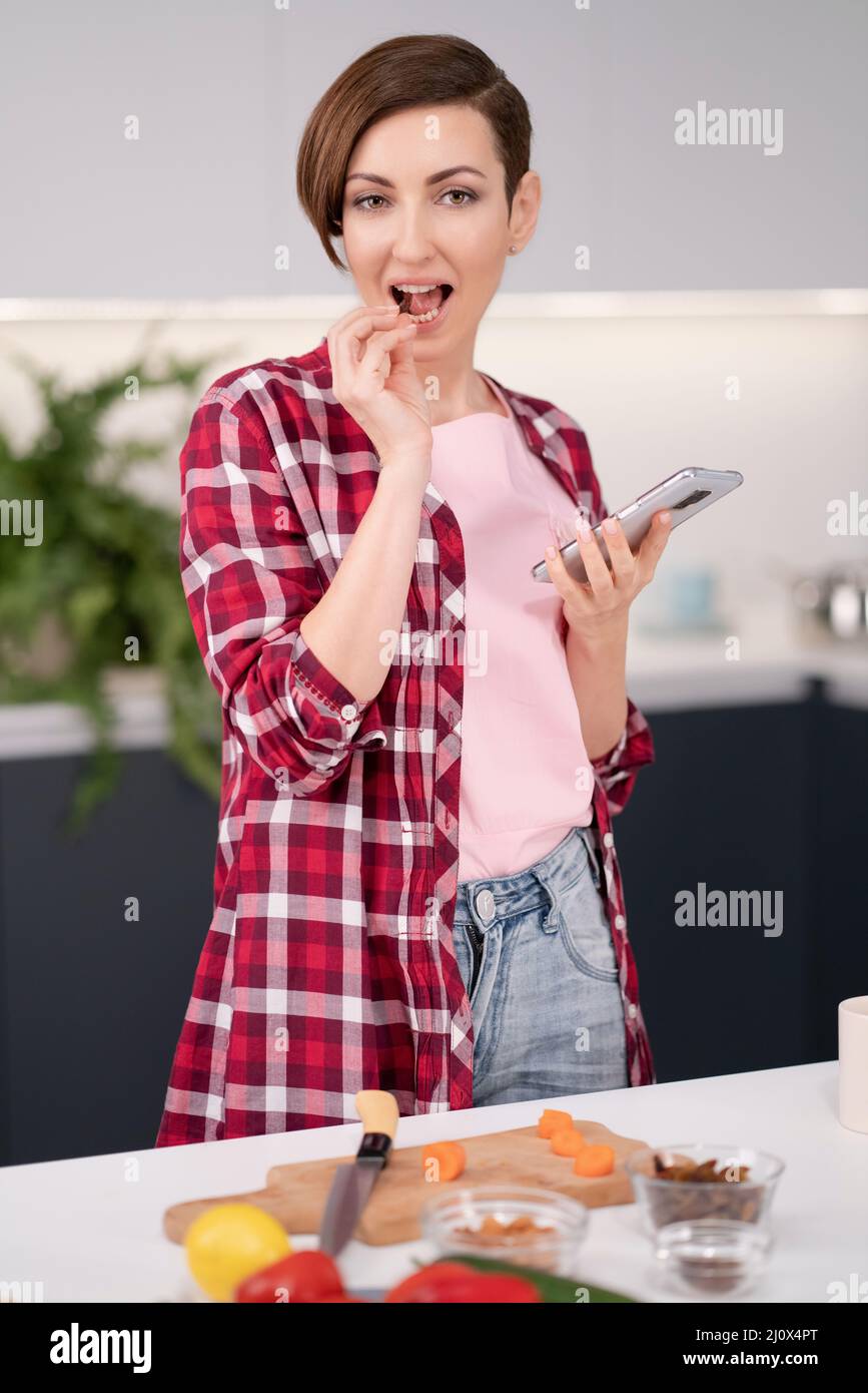 Beautiful woman hold smartphone in hands reading messages or social media with short hair and red shirt makes fresh food at home Stock Photo