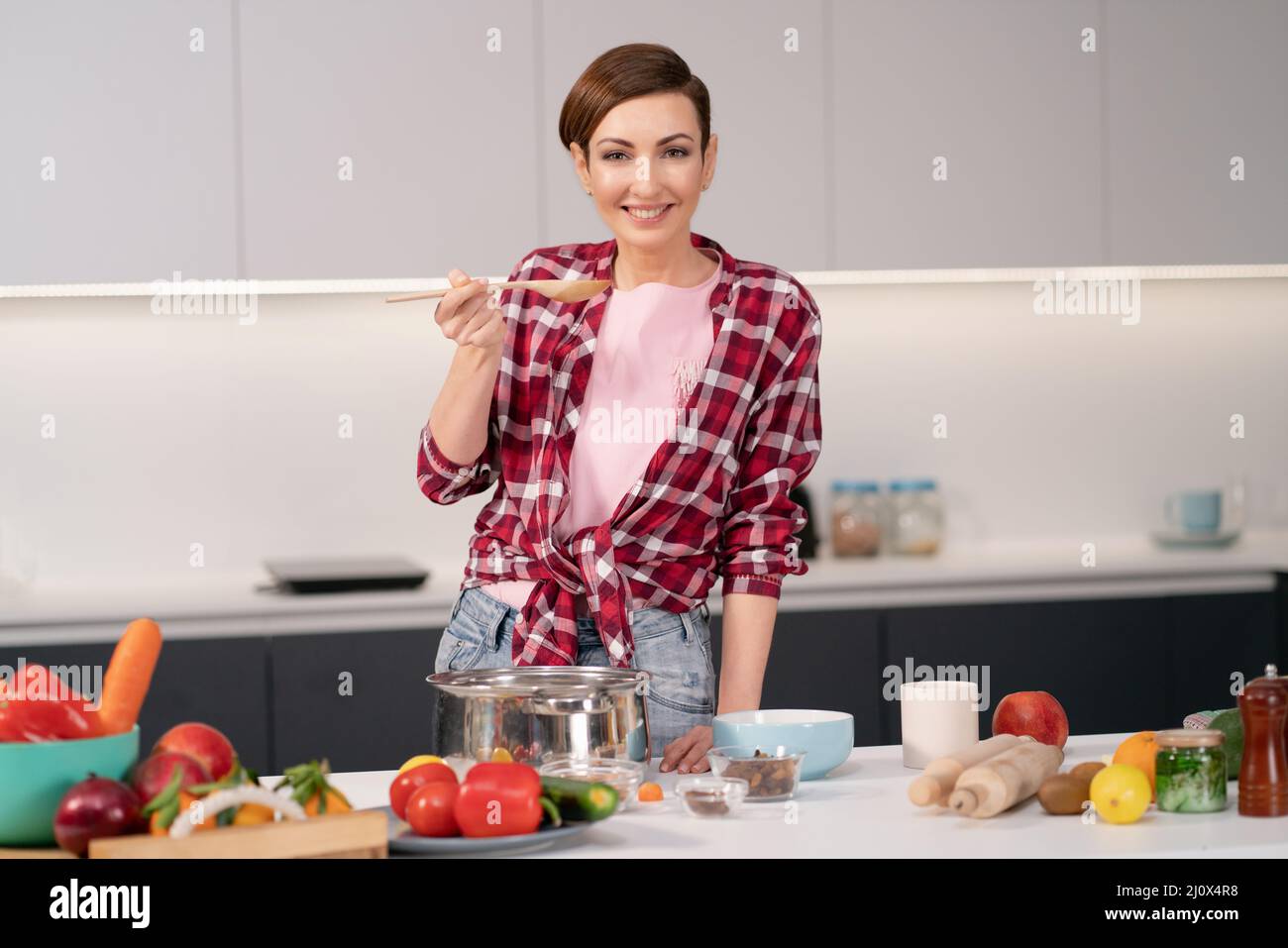 Cheerful housewife in red plaid shirt cooking soup on modern kitchen with lots fresh vegetables on the table in front of her. Sm Stock Photo