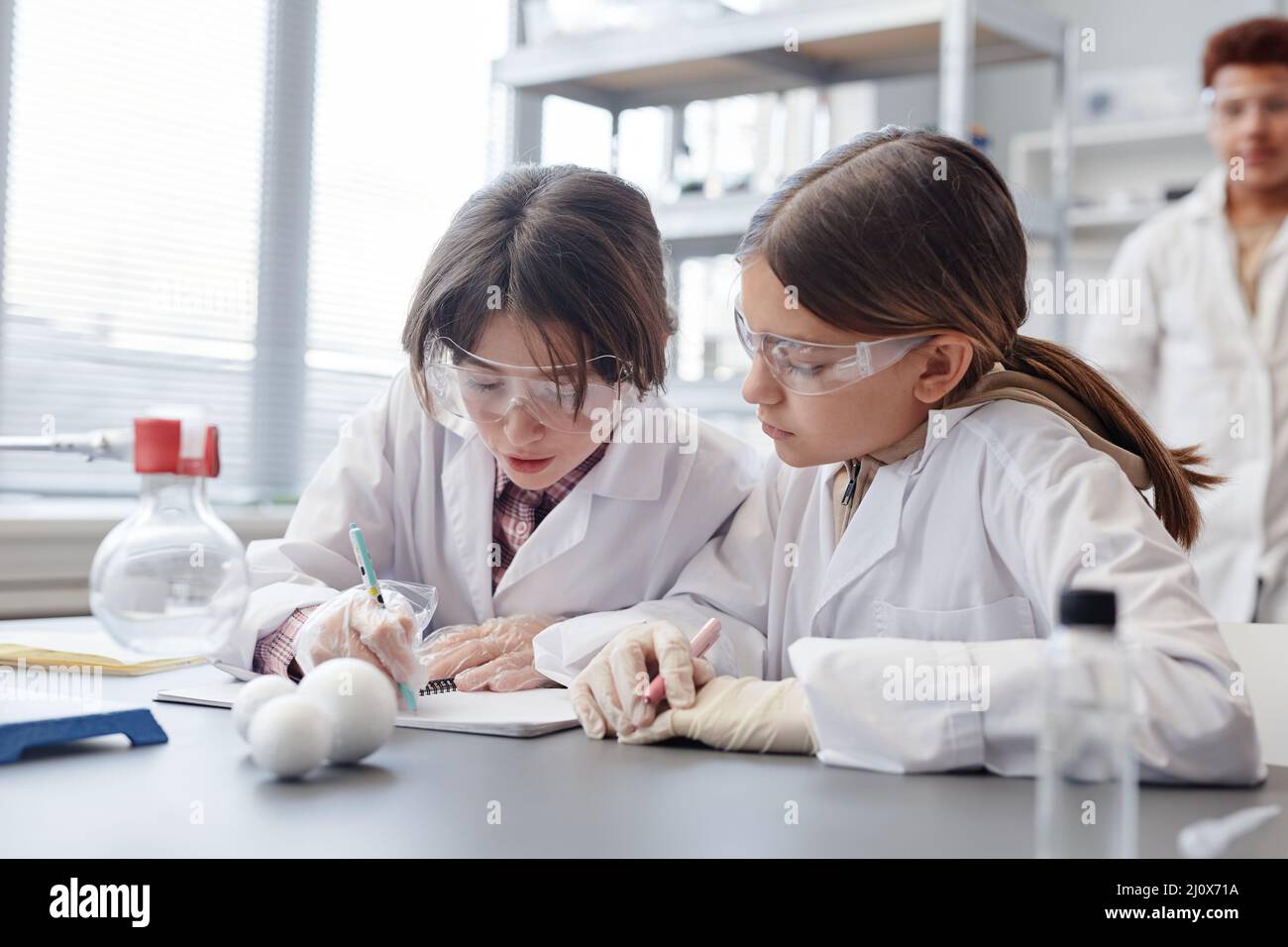 Portrait of two girls taking notes in notebook while doing science experiment in chemistry lab at school Stock Photo