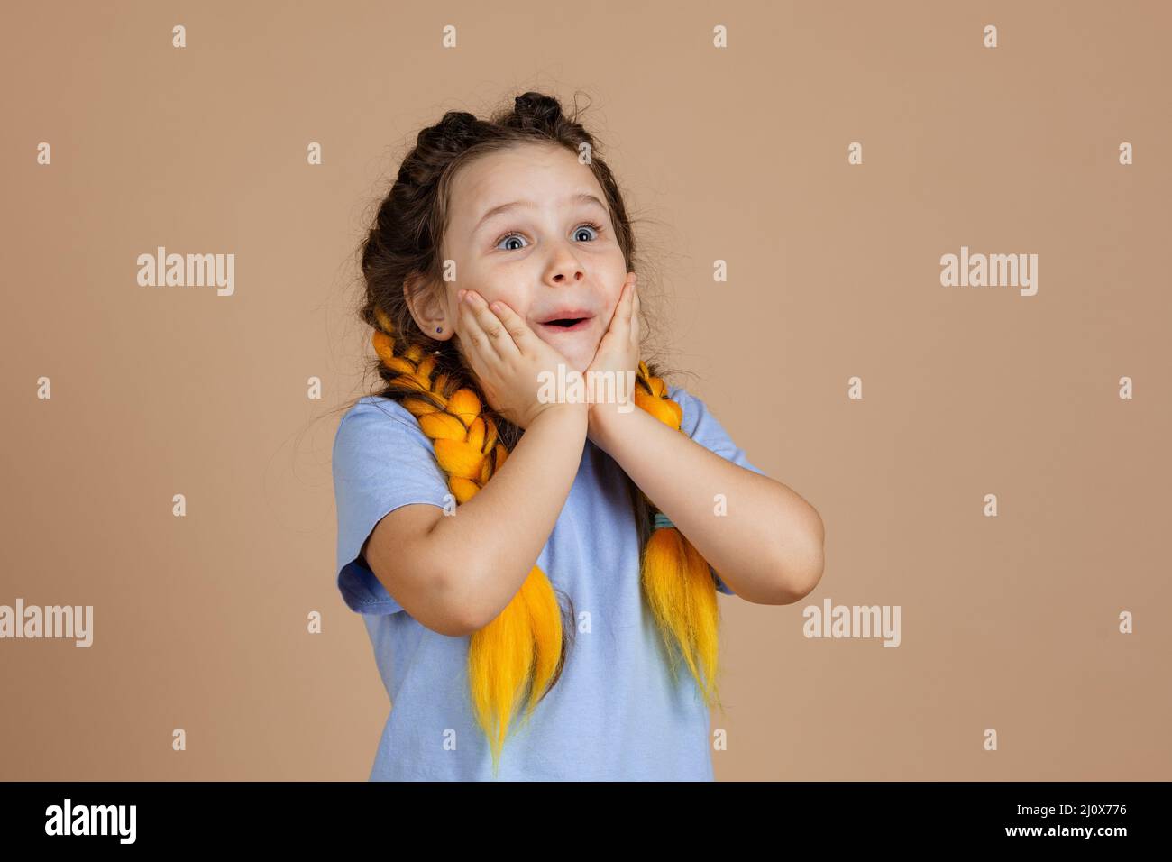 Surprised shocked girl touching face with hands looking up with shining eyes with yellow kanekalon braids on head on beige background wearing blue t Stock Photo