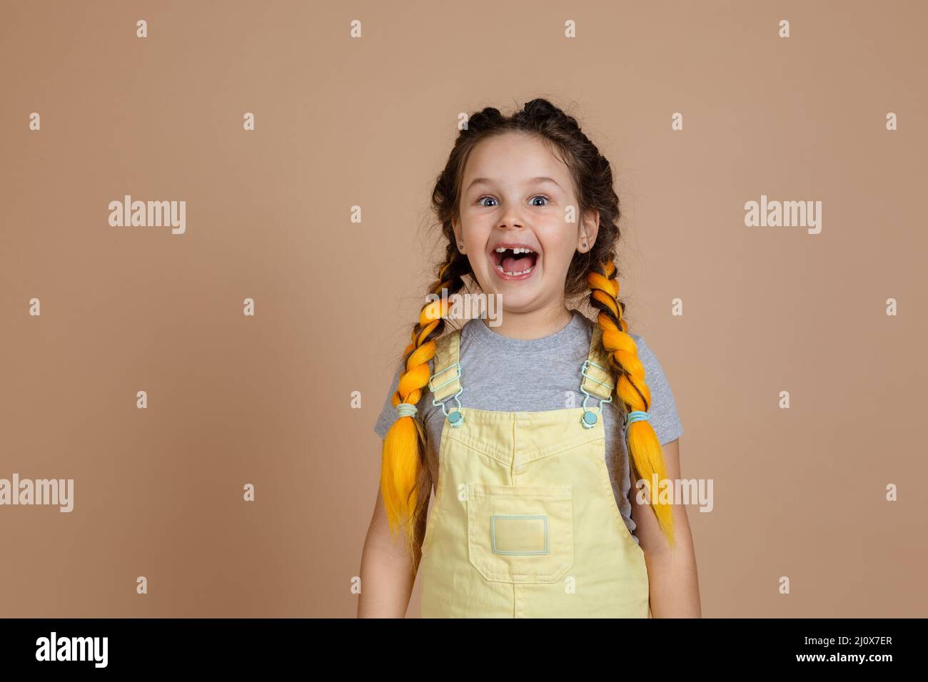 Playful excited small female with yellow kanekalon pigtails, smiling with opened mouth with missing tooth wearing yellow jumpsuit and gray t-shirt on Stock Photo