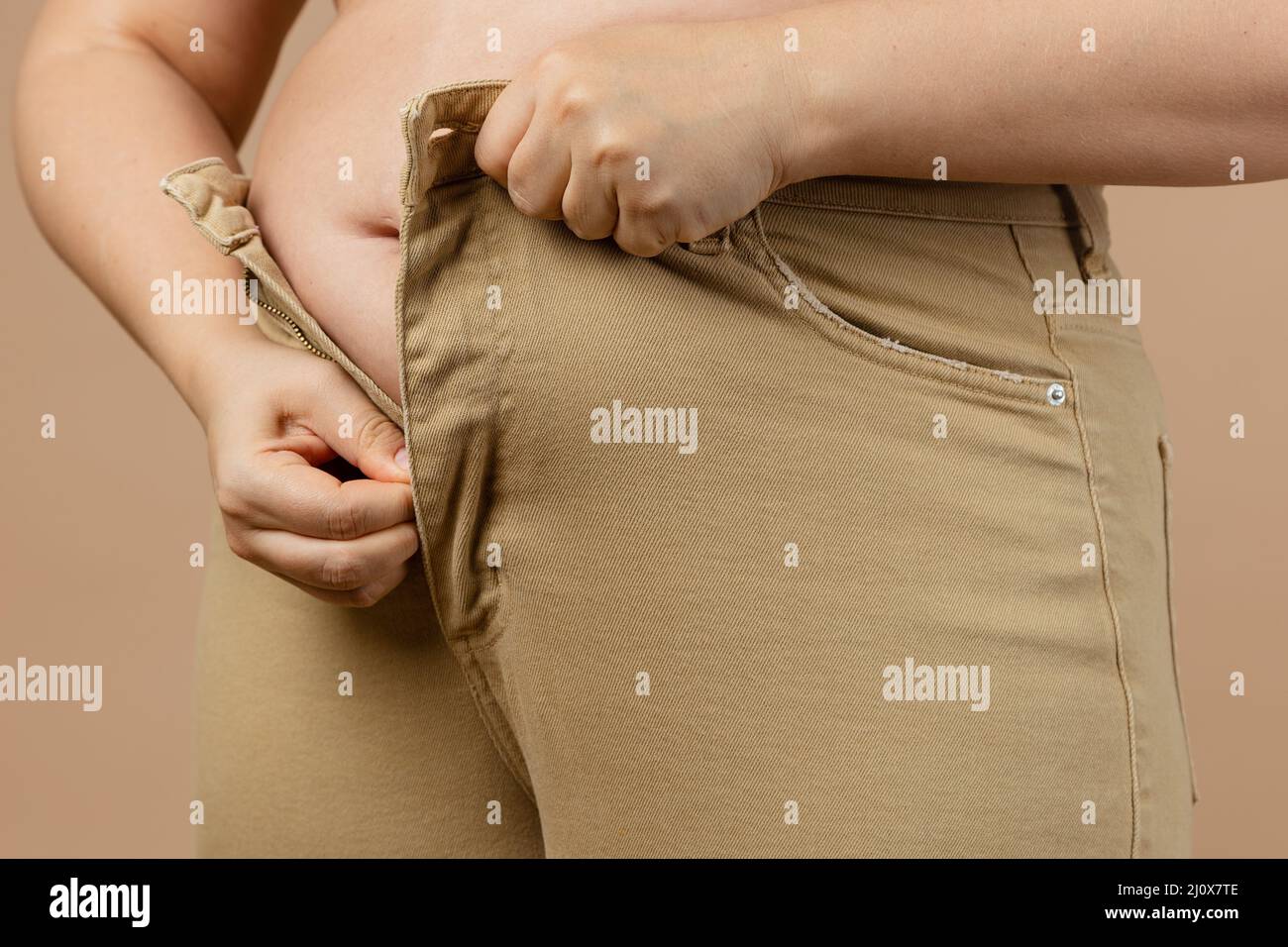 Woman big tummy not allowing her to wear jeans on beige background. Visceral fat. Body positive. Sudden weight gain. Tight little clothes. Need for Stock Photo