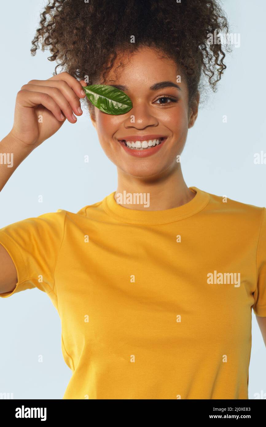 Natural beauty. Happy Afro american girl in yellow tshirt with curly hair covering one eye with green leaf. Stock Photo