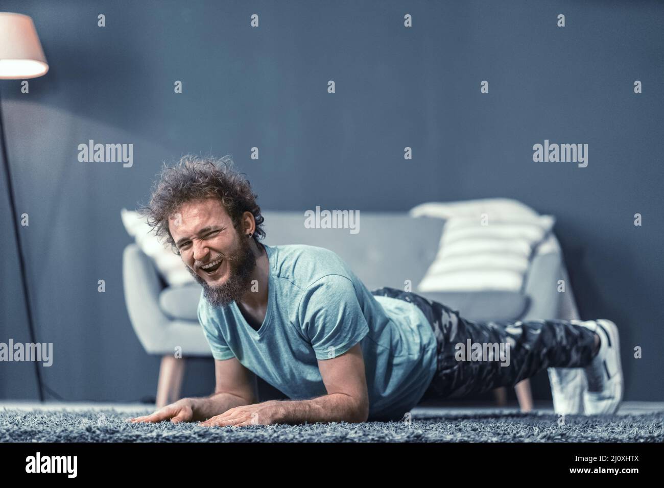 Handsome man doing push up or abs core plank exercise at home laughing looking at camera. Funny curly hair man with a mess beard Stock Photo