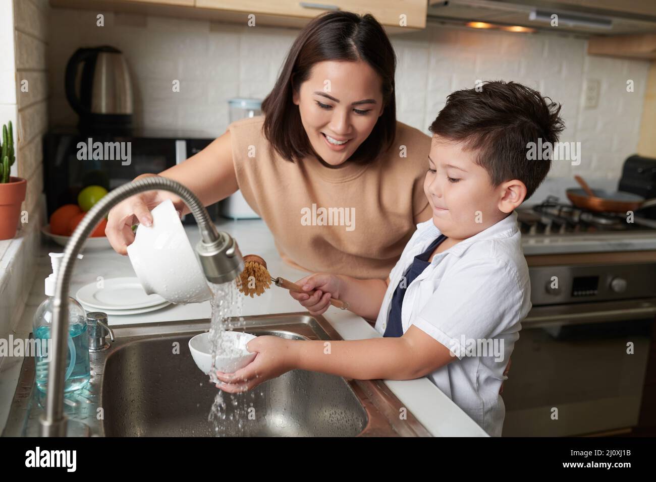 Smiling mother helping son to rinse dishes under tap water after