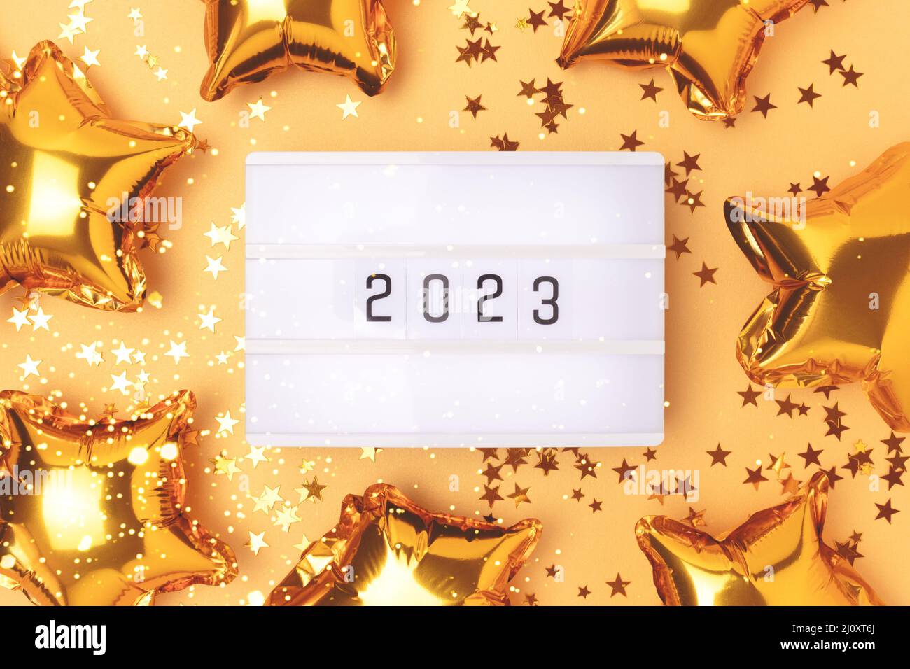Lightbox with 2023 numbers on a gold colored background. Frame made of stars confetti and foil balloons. Stock Photo