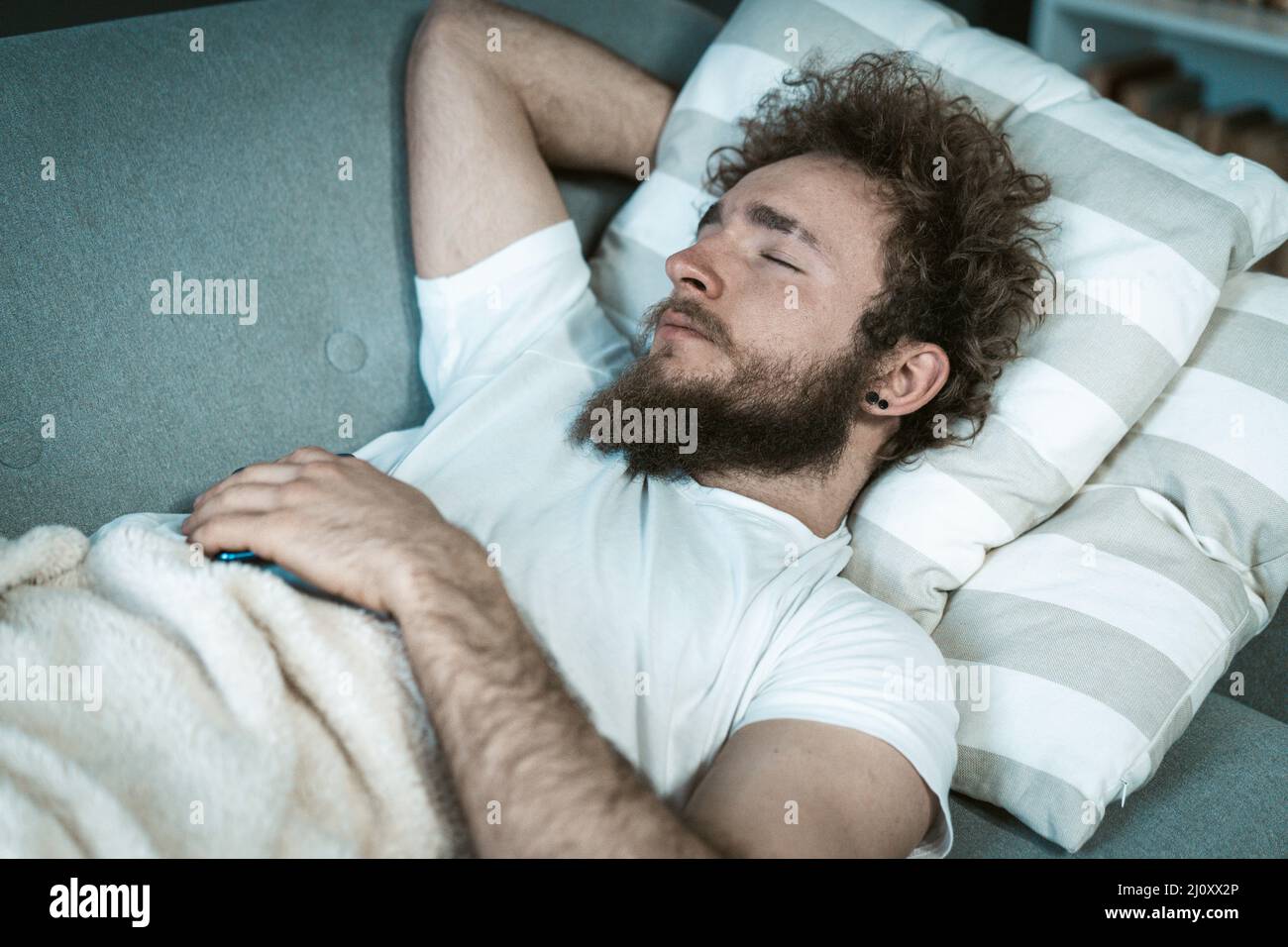 Relaxed sleeping handsome man with curly hair mid day resting on the couch or sofa enjoying weekend or quarantine days at home. Stock Photo