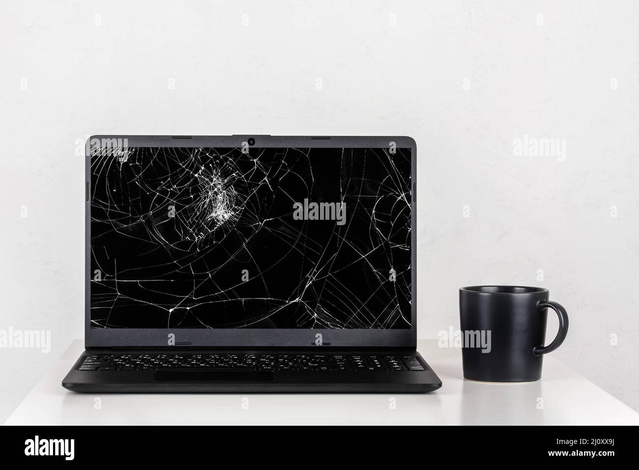 A laptop with a broken screen and a black mug of tea on a table against a white wall Stock Photo