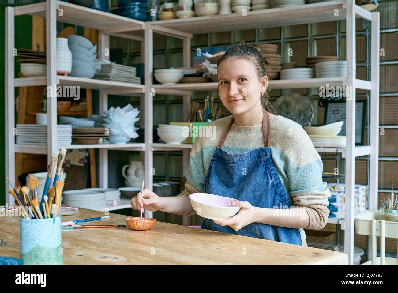 Skilled young woman in apron standing at table and drawing on ceramic bowl in pottery workshop Stock Photo