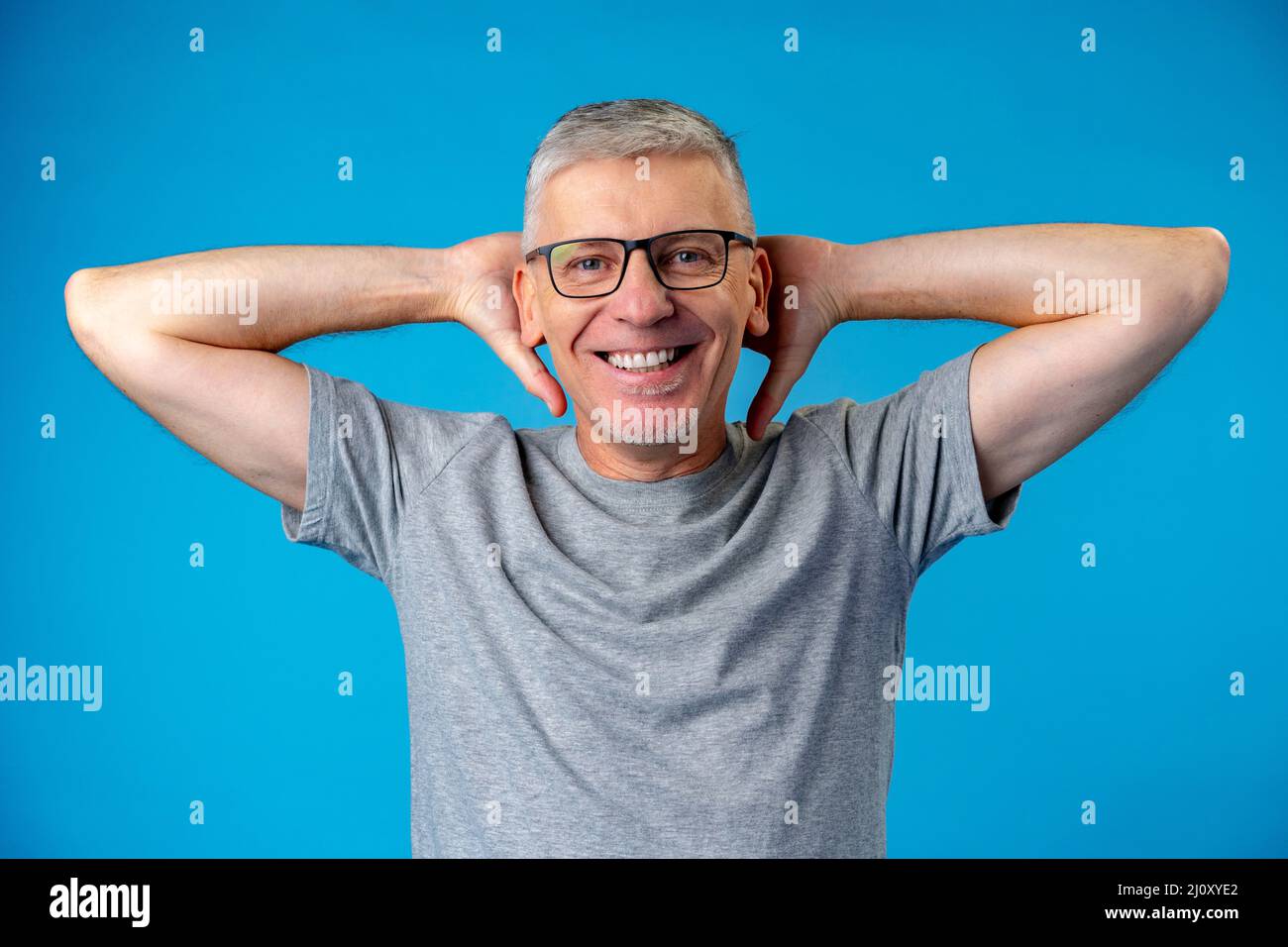 Middle aged man standing on blue backgorund with hands behind head Stock Photo