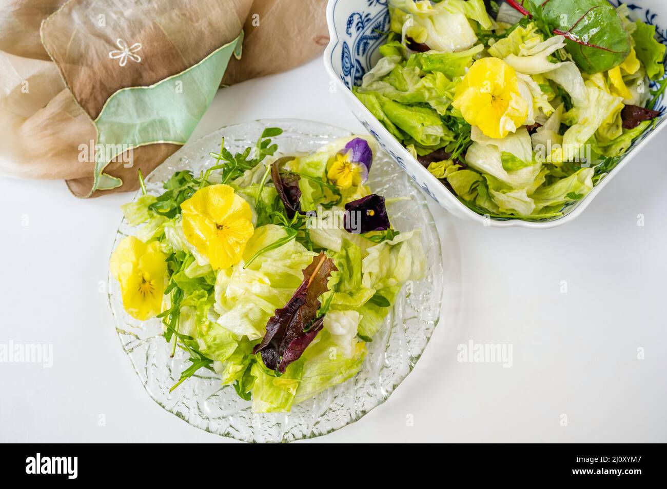 Fresh green salad (lettuce,rucola,red chard) with edible pansy flower on glass plate and in bowl, scarf on white background, closeup. Stock Photo