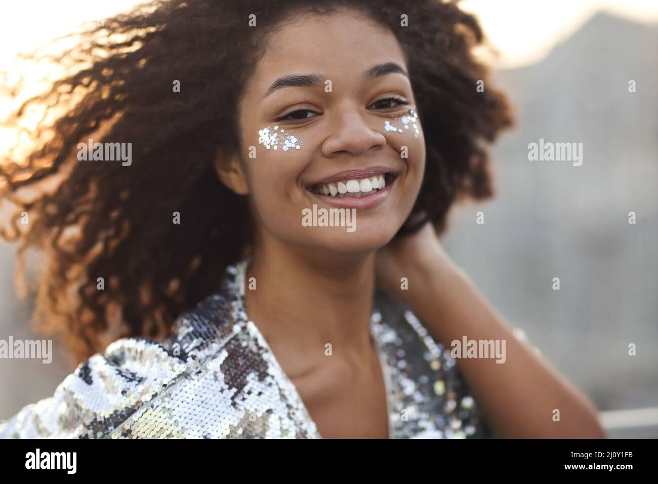 Close up portrait of overjoyed charming african american woman with curly hair and glitter on cheekbones smiling happily at came Stock Photo