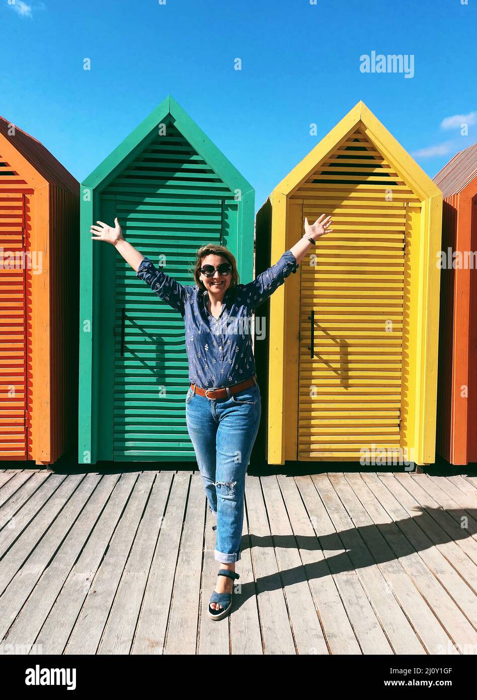 Happy young woman raising hands and smiling while standing against colourful wooden beach cabins Stock Photo