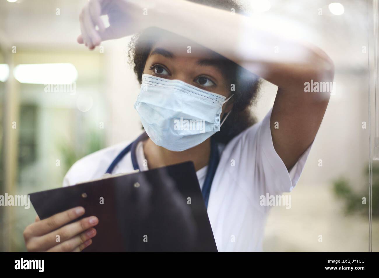 Young tired overworked african american medical worker in protective face mask looking out of window Stock Photo