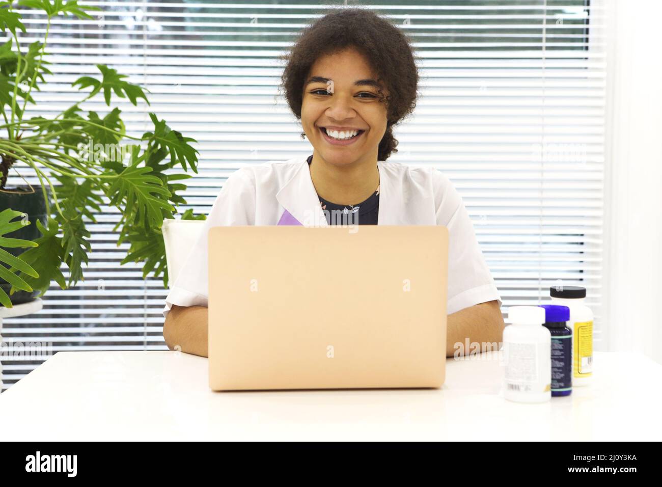 Young happy African-American female doctor sitting at workplace looking to the camera smiling while working on laptop in modern Stock Photo