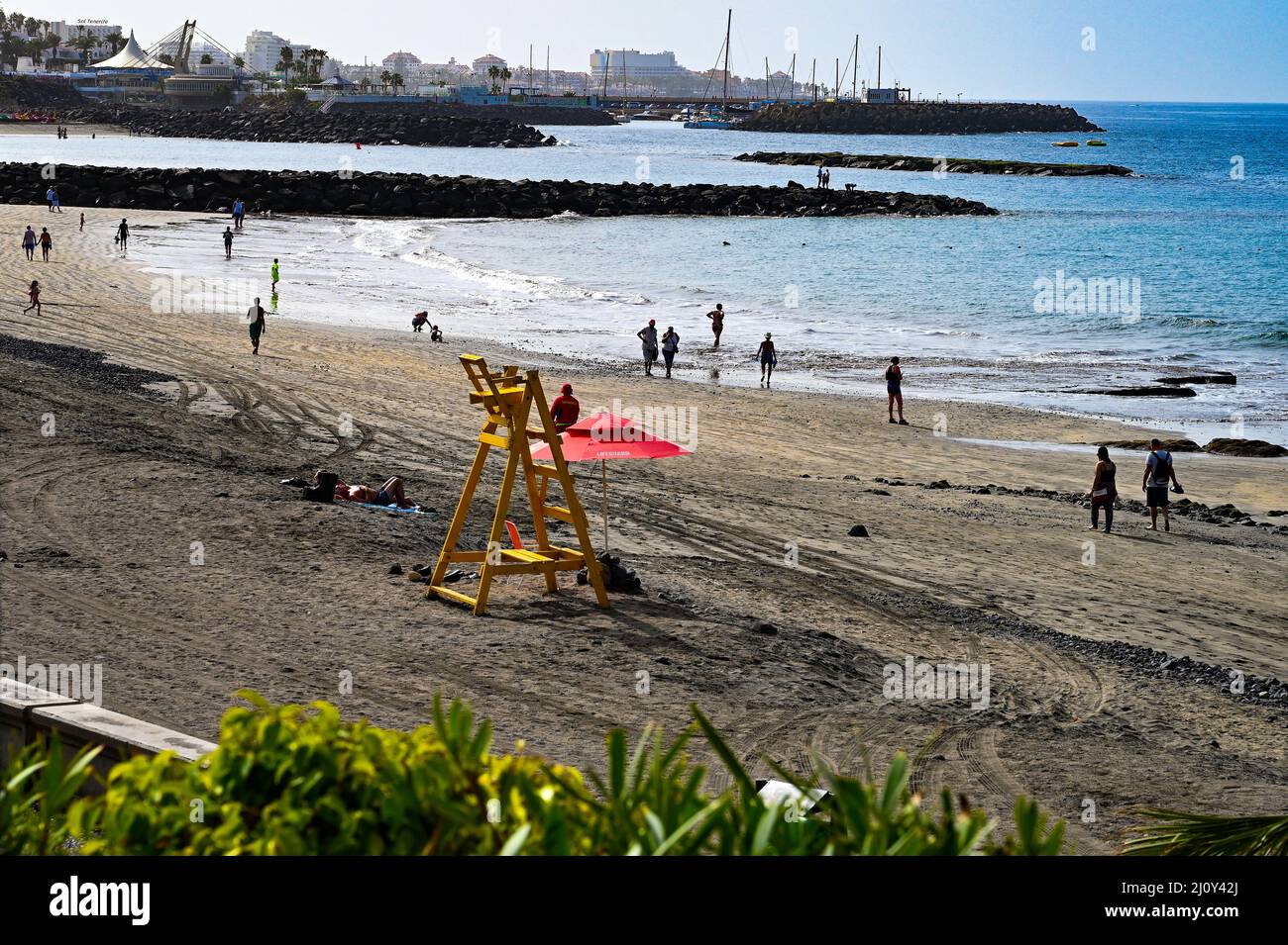 An empty yellow life savers, life guards chair on the volcanic beach with holiday makers enjoying the sunshine in Costa Adeje, Tenerife, Spain Stock Photo