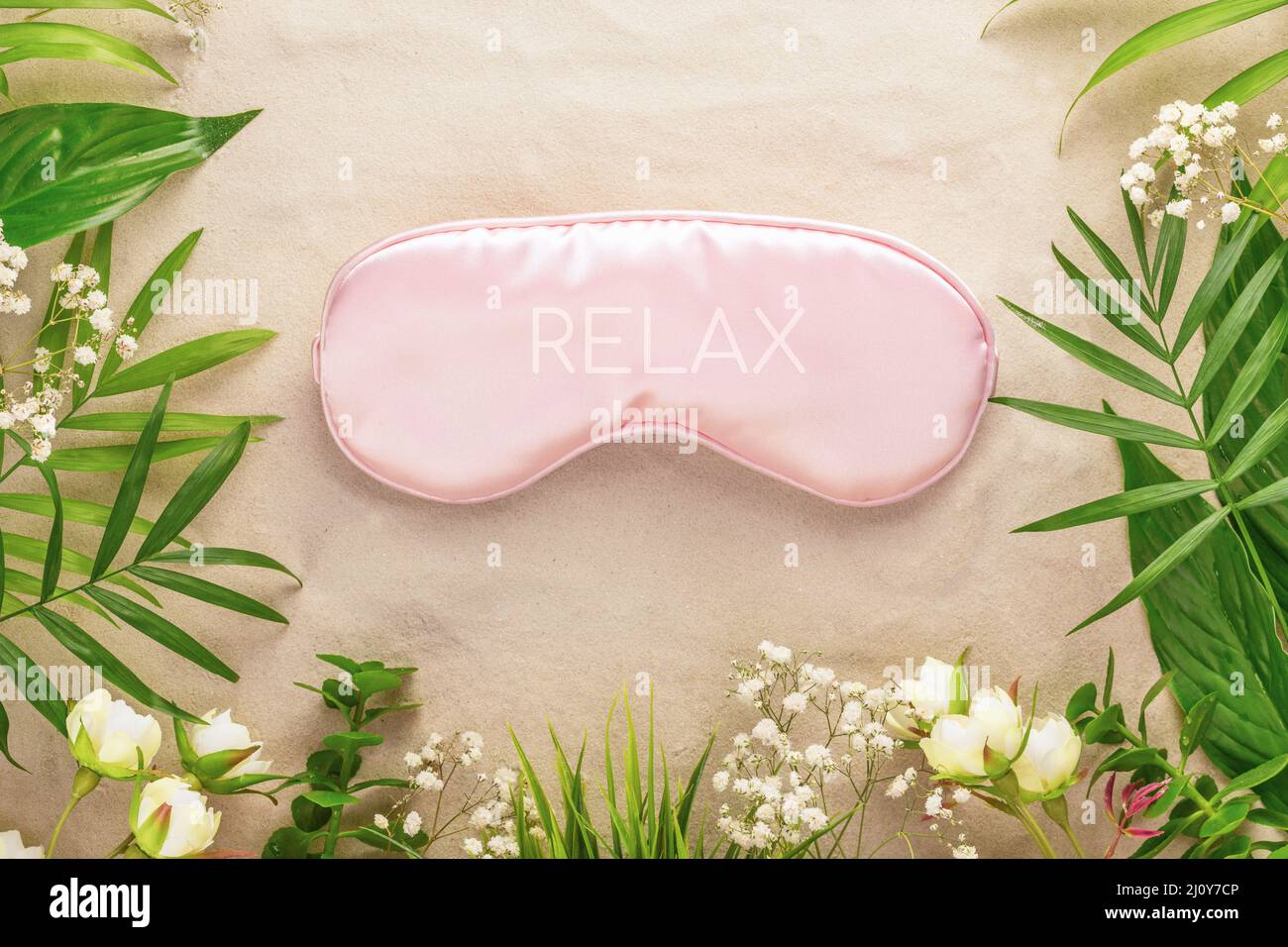 Relax concept. Wellbeing composition with pink mask for sleep, tropic plant and flowers on a sand background. Insomnia or depression treatment and men Stock Photo