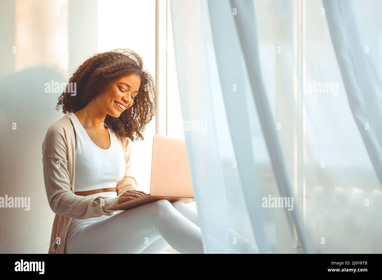Happy african american young woman freelancer on windowsill with laptop on laps, working from home Stock Photo