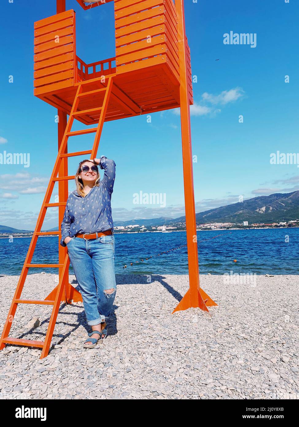 Happy smiling woman in casual clothes standing on beach on sunny summer day near lifeguard stand Stock Photo