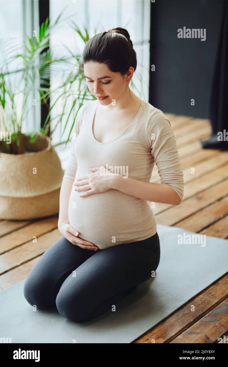 Meditation during pregnancy. Young calm tranquil pregnant woman doing yoga at home, meditating and practicing mindfulness, expec Stock Photo