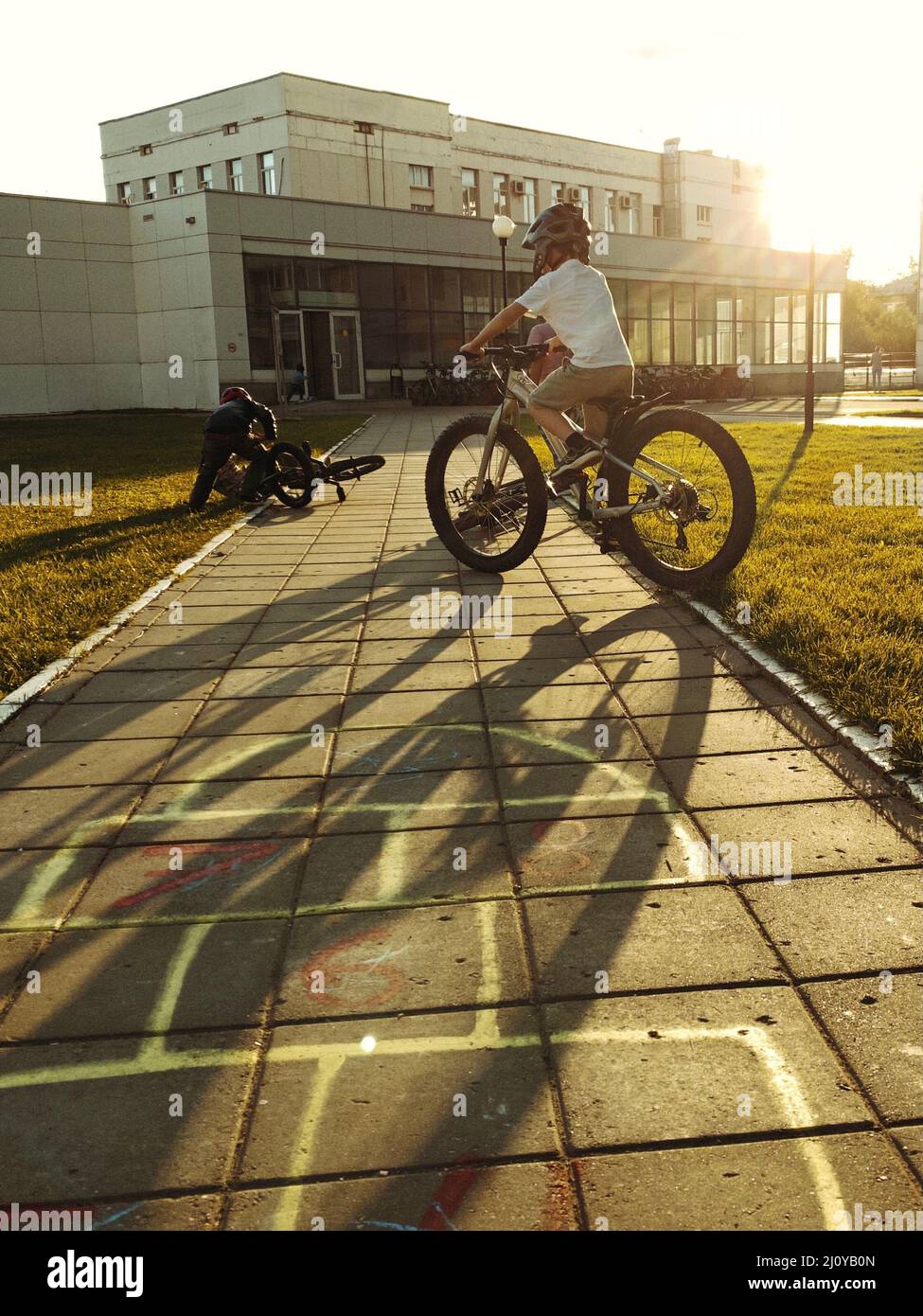 On warm autumn evening, boy in big helmet rides bicycle with huge wheels on road. Boy picks up fallen bike. At sunset there are big shadows from bike Stock Photo