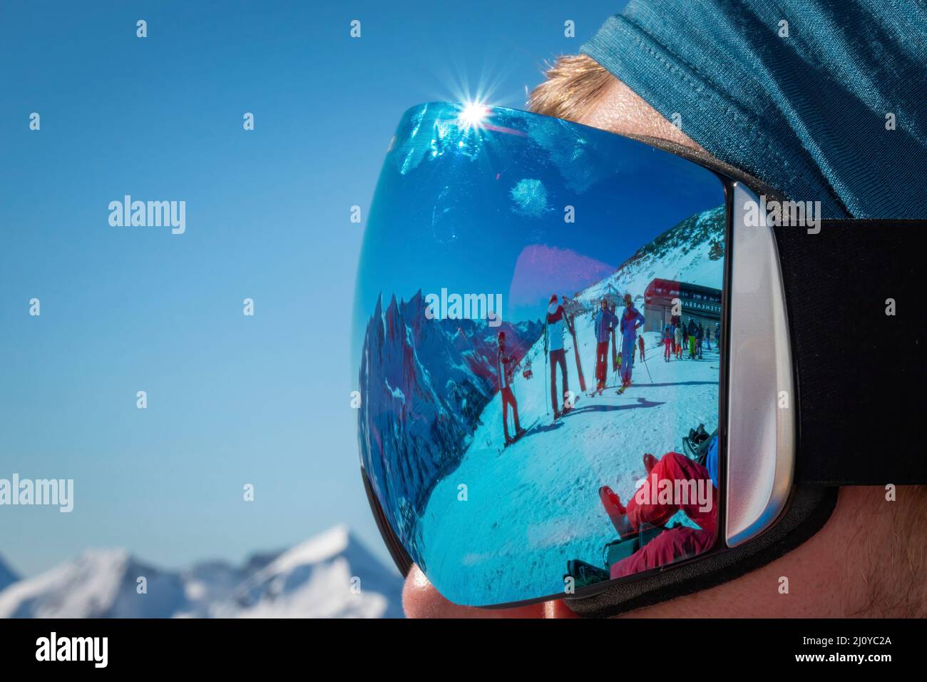 Close-up of man wearing ski goggles with reflecting mountains and skiers in winter against blue sky Stock Photo