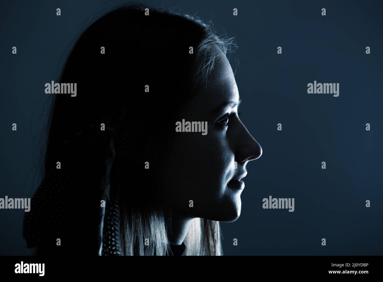 Close-up woman with feathers in her hairs dark profile portrait. Model face partly illuminated by blue light imitating television or moon light. Selec Stock Photo