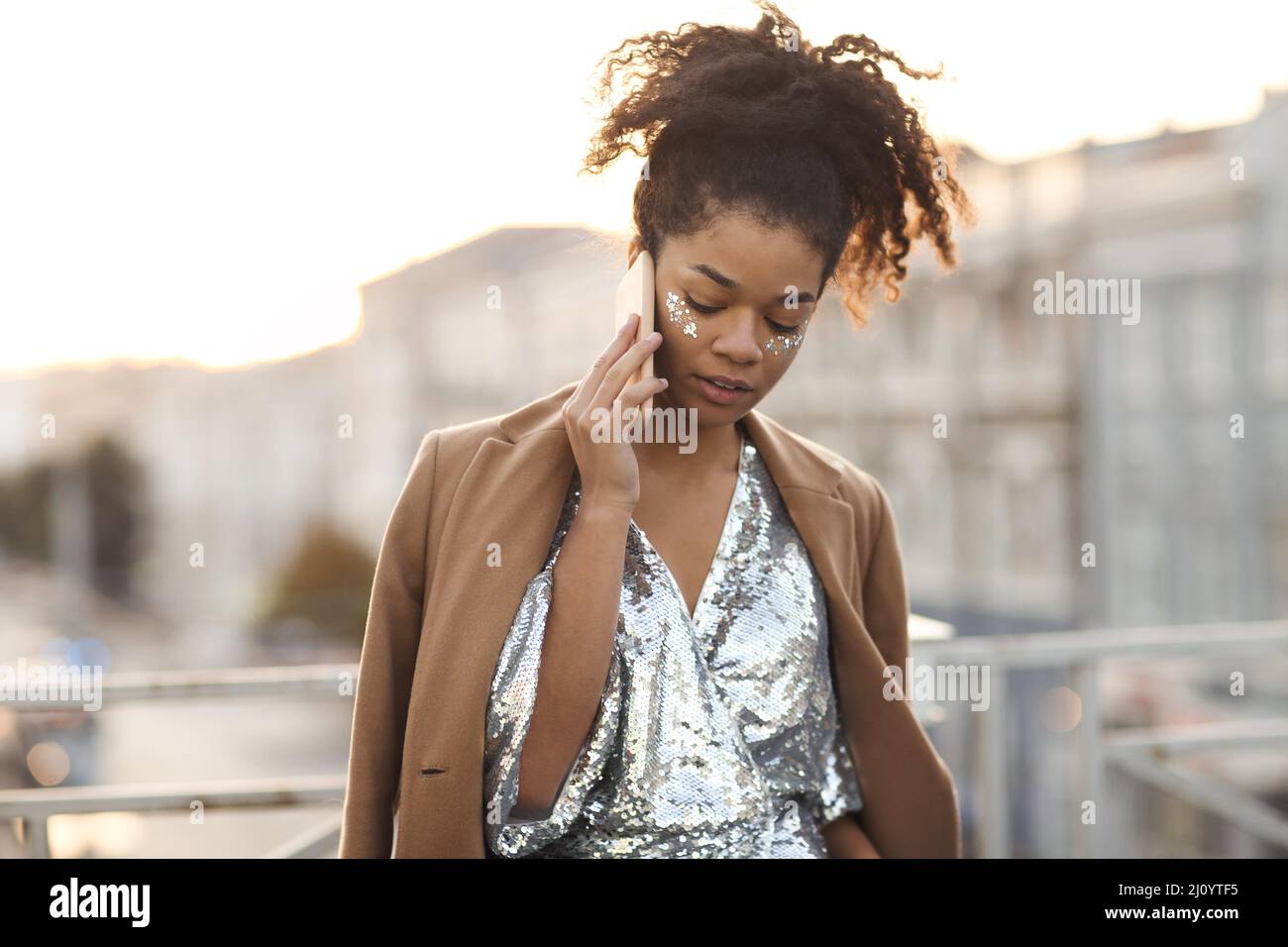 African american woman with glitter on cheekbones in sequin dress talking on mobile phone at rooftop Stock Photo