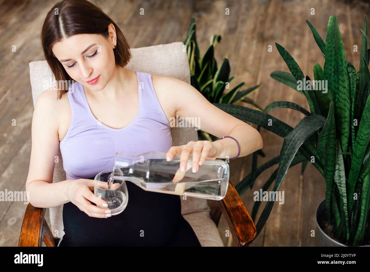 Hydration during pregnancy. Young pregnanat woman in t-shirt sitting on bed at home, drinking clean mineral water in morning to prevent dehydration. e Stock Photo