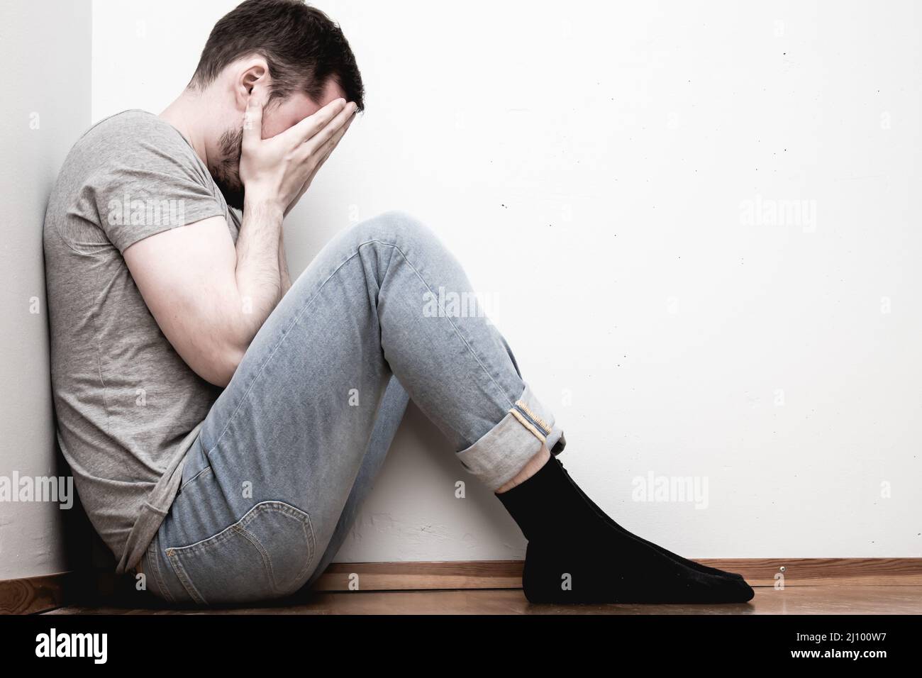 Depressed male sitting in the corner of a room hiding his face with two ...