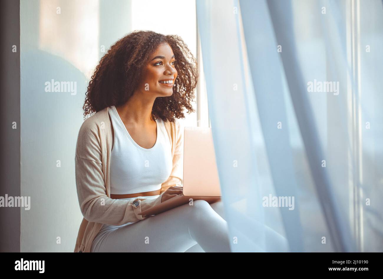 Happy african american young woman freelancer on windowsill with laptop on laps, working from home Stock Photo
