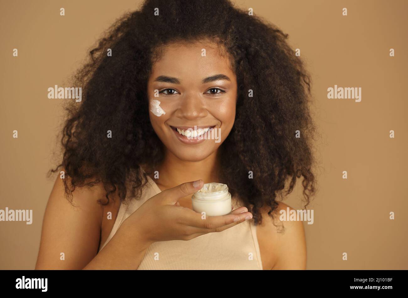 Close up shot of smiling dark skinned woman with curly afro hair applying face cream Stock Photo