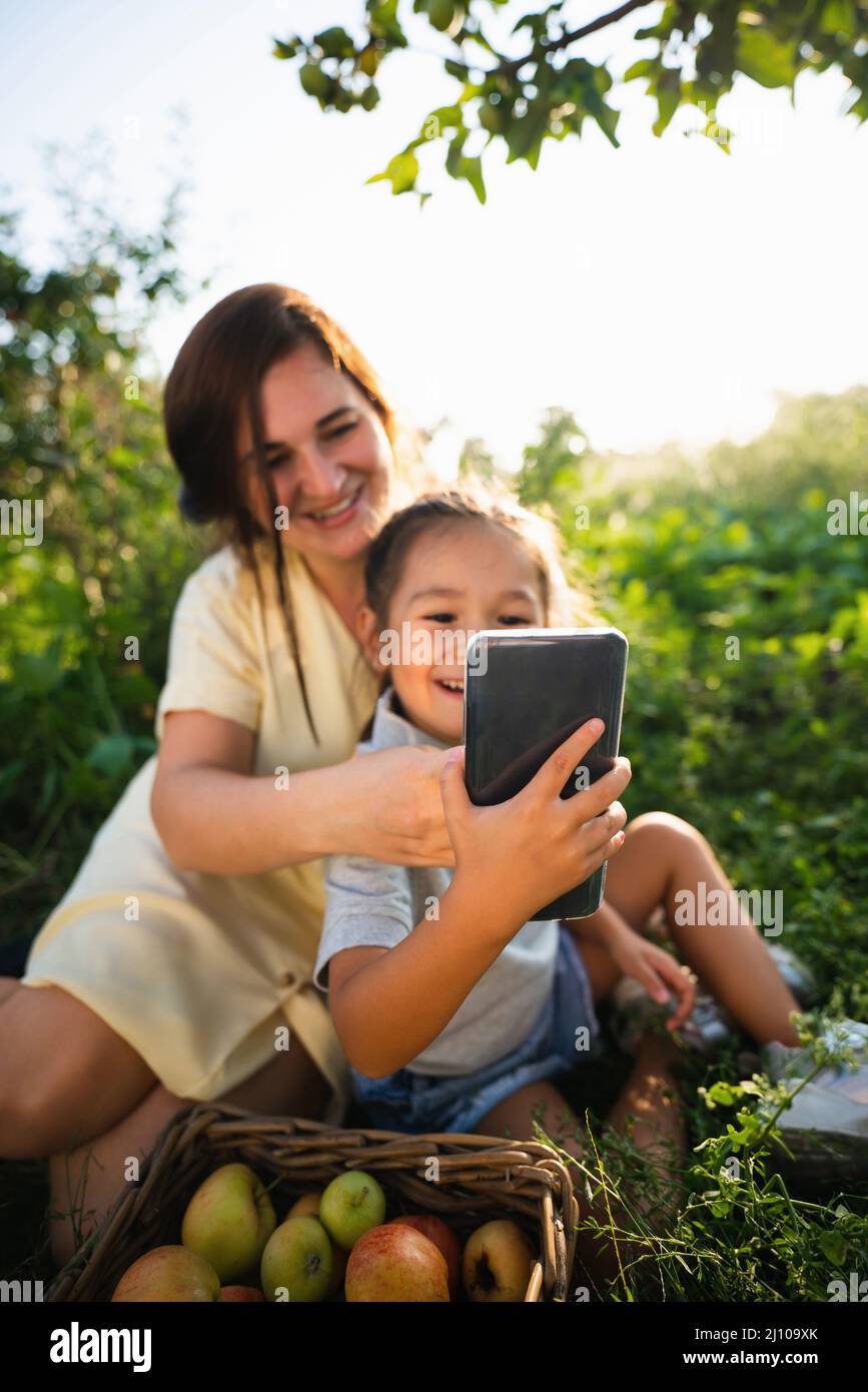 Caucasian mother and her Asian daughter take a selfie in the orchard in summer  Stock Photo