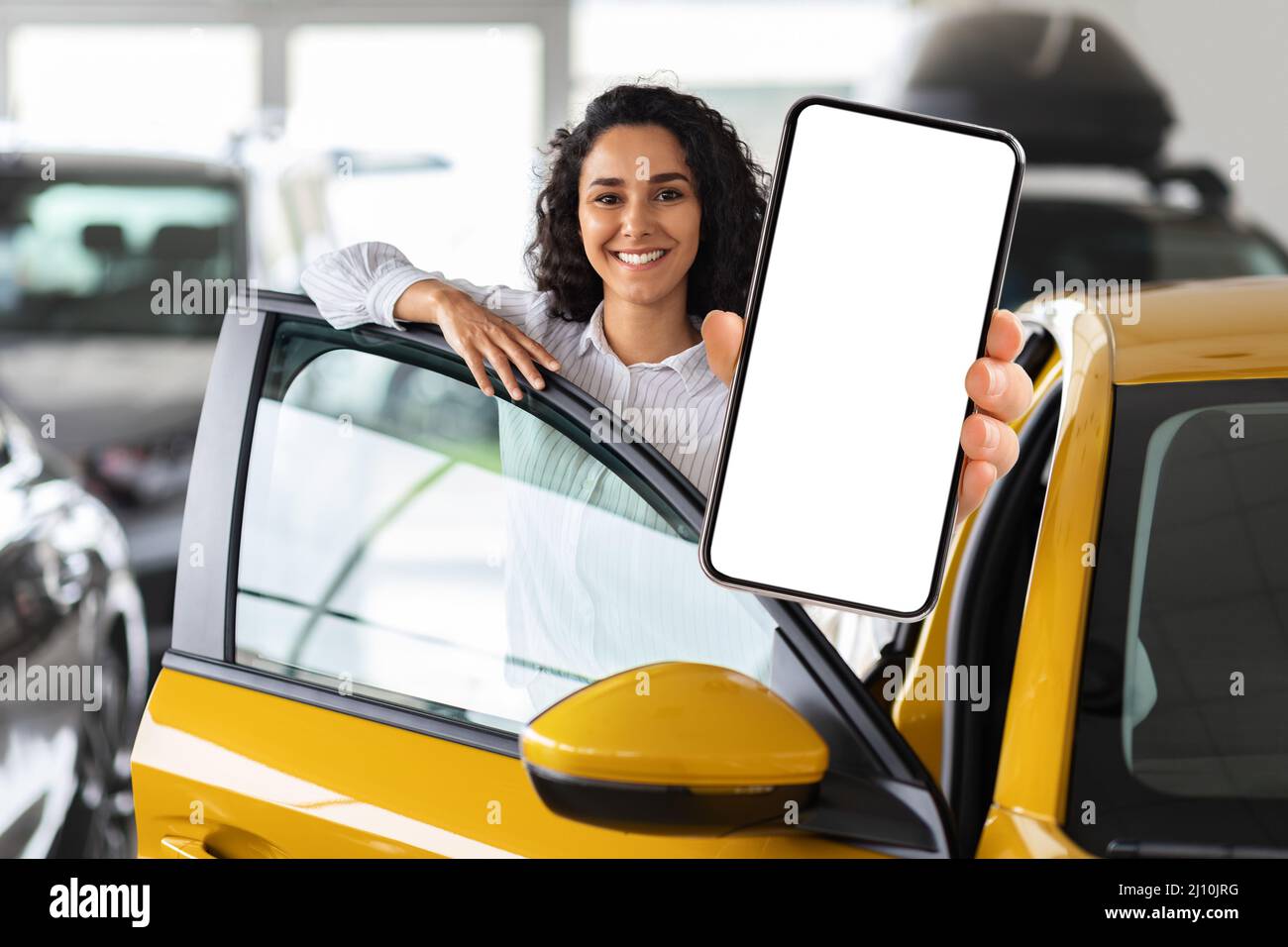 Positive middle eastern woman standing by car, showing smartphone Stock Photo