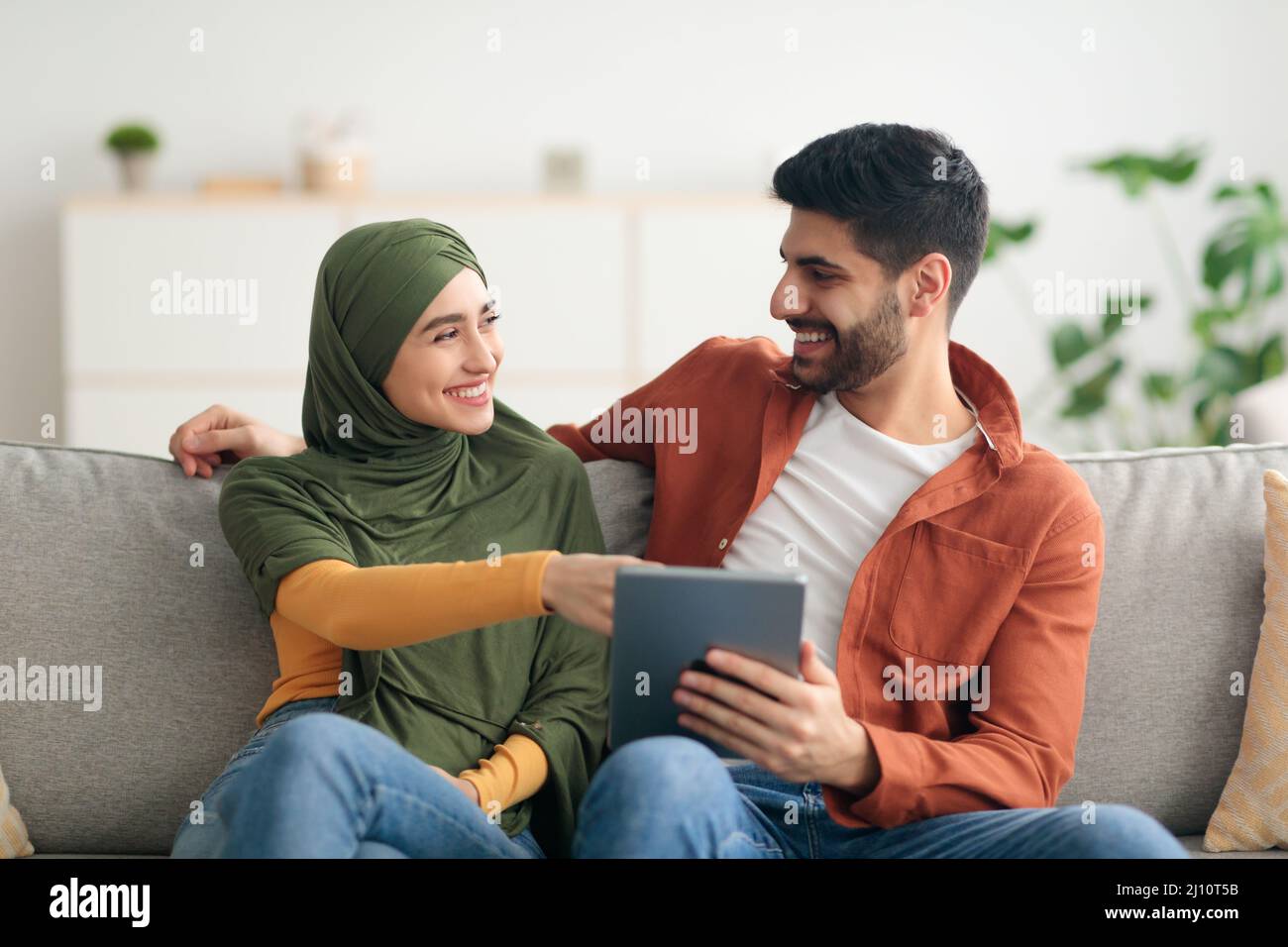 Cheerful Arabic Spouses Using Tablet Computer Sitting On Couch Indoor Stock Photo