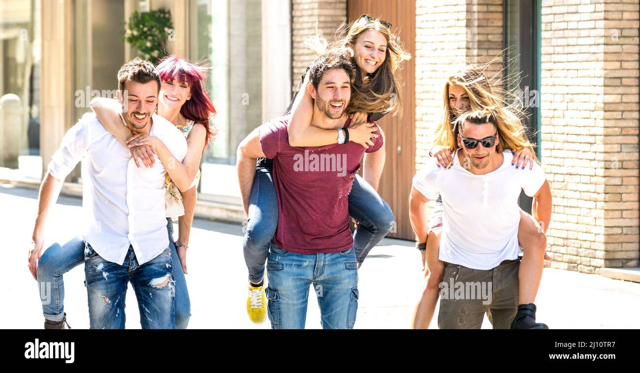 Happy millennial friends couples walking in city center on piggyback move - Happy guys and girls having fun around old town streets - University stude Stock Photo