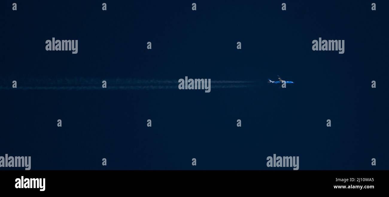 A commercial airliner at high altitude with contrails passes through a deep blue sky at sunset. Stock Photo