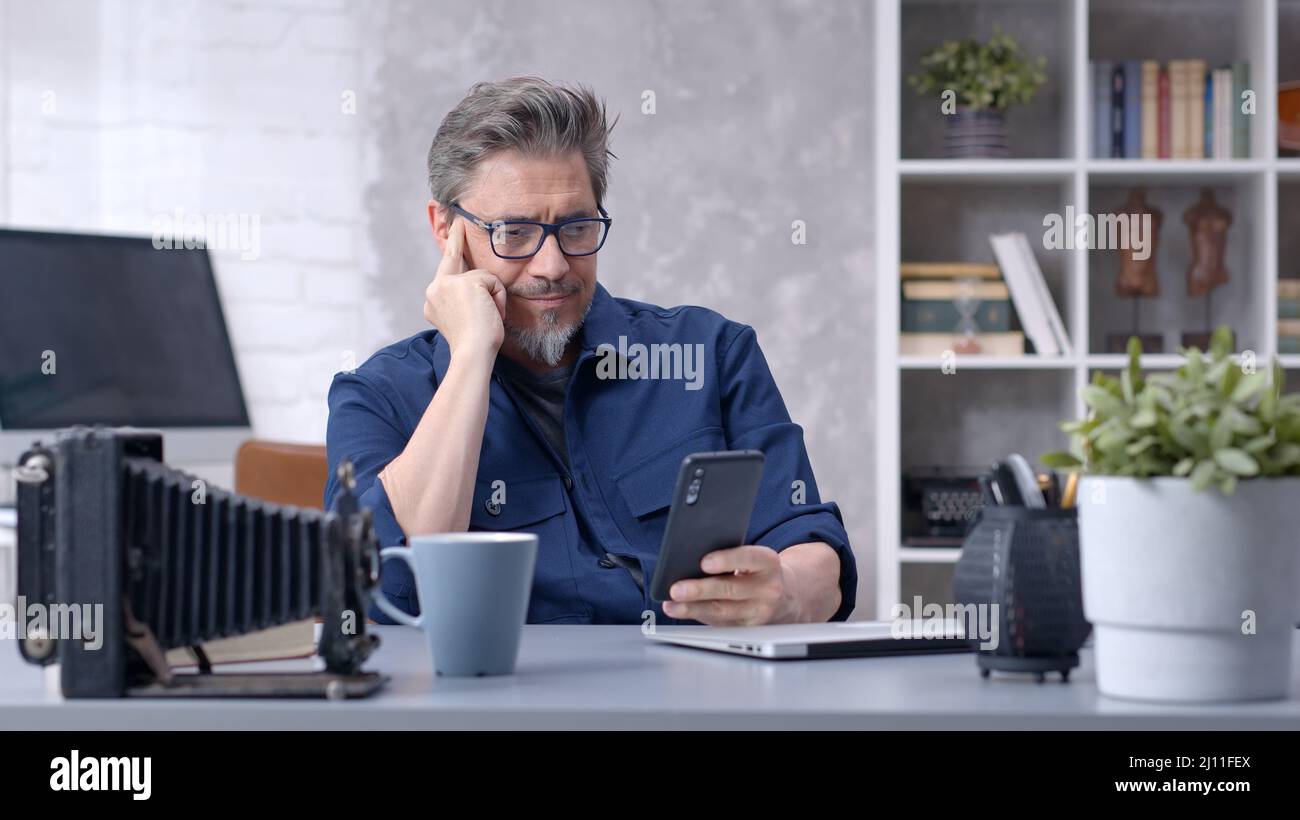 Bearded man working online with laptop computer and smart phone at home sitting at desk. Businessman in home office. Portrait of mature age, middle age, mid adult man in 50s. Stock Photo