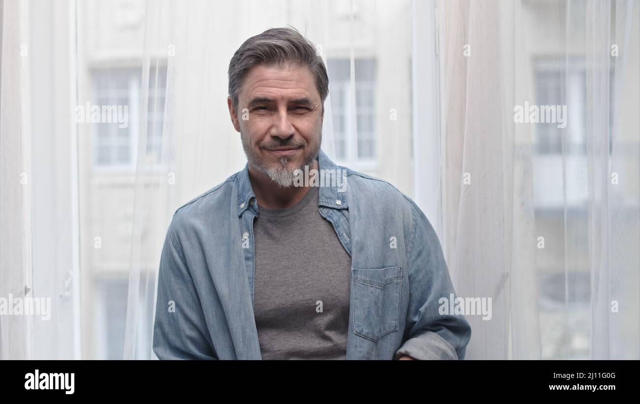 Portrait of older grey haired man standing in window at home, smiling. Stock Photo